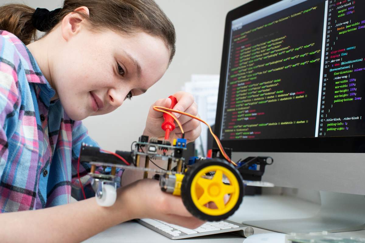 Mädchen schraubt an programmierbarem Auto an einem Schreibtisch mit Codezeilen auf dem Bildschirm